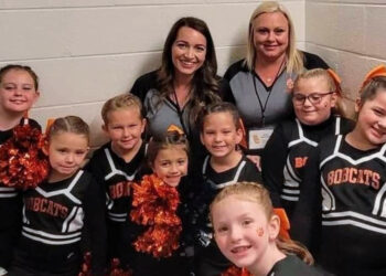 The cheerleaders and coaches stand against a white wall