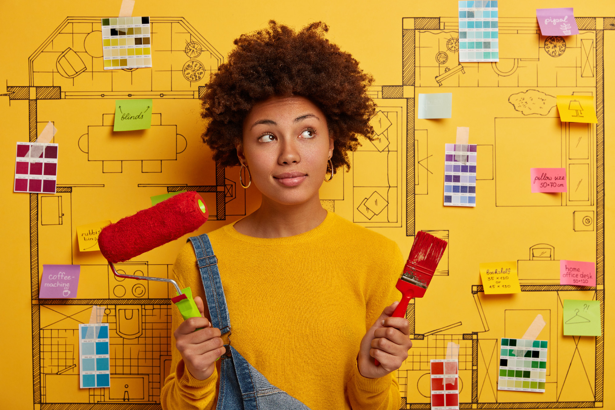 Contempative curly female interior designer with paint roller and brush, does repairing in room, thinks how to improve design project of new apartment. Repair, designing and flat renewal concept