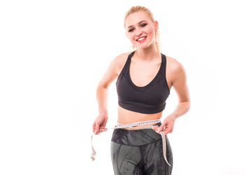 Attractive female bodybuilder measuring her waist with measurement tape. Photo of young woman in sportswear on white background. Strength and motivation