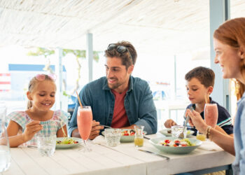 Cheerful family in restaurant enjoying lunch together in a patio. Mother and father observing daughter while eating food with son busy in eating. Smiling family with two children eating brunch together at kiosk during summer vacation.